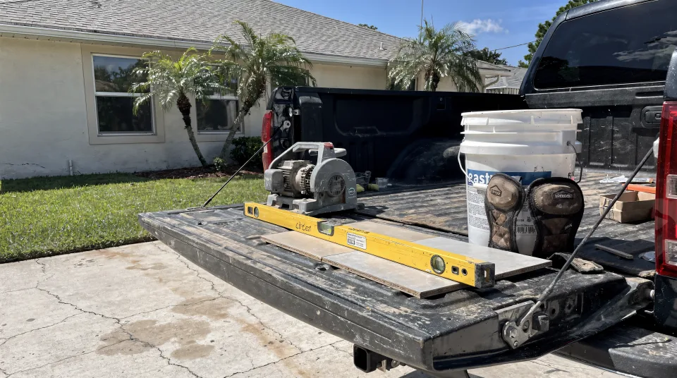 Work truck tailgate in a Florida driveway with a tile saw, a level, a bucket and a box of porcelain tile laid out before a job