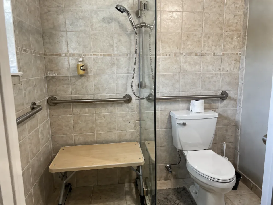 Finished accessible shower with two grab bars, a hand-held shower on a slide bar and a fold-down bench