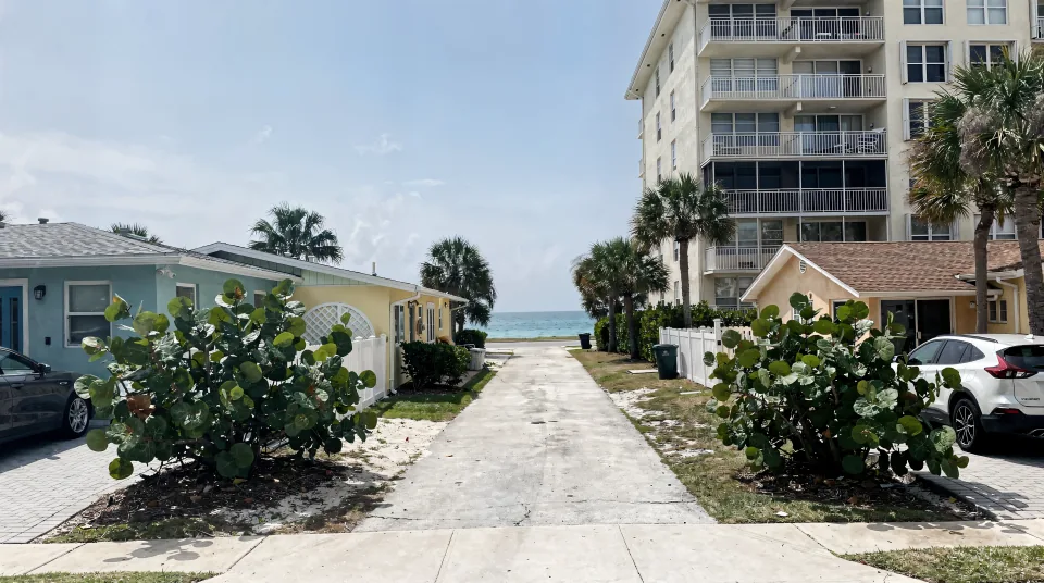 Pompano Beach street near the water with a mid-rise condo beside older single-story cottages, sea grapes and a strip of ocean