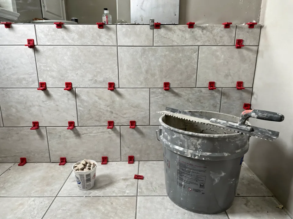 Large-format porcelain tile half set on a shower wall with red leveling clips and a bucket of thinset