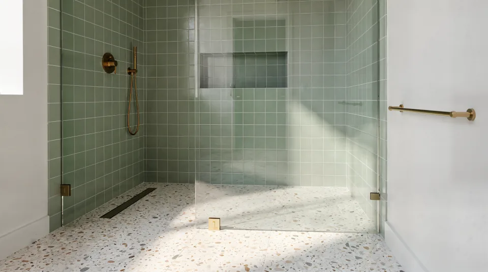 Finished bathroom with a curbless walk-in shower, soft green ceramic wall tile, a terrazzo-look floor, brass fixtures and a frameless panel (a rendering of the finished look, not a job photo)