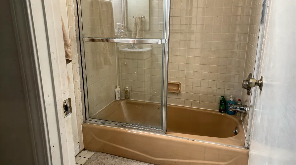Dated 1980s Florida bathroom with an almond bathtub, a sliding glass door and beige tile surround seen from the doorway