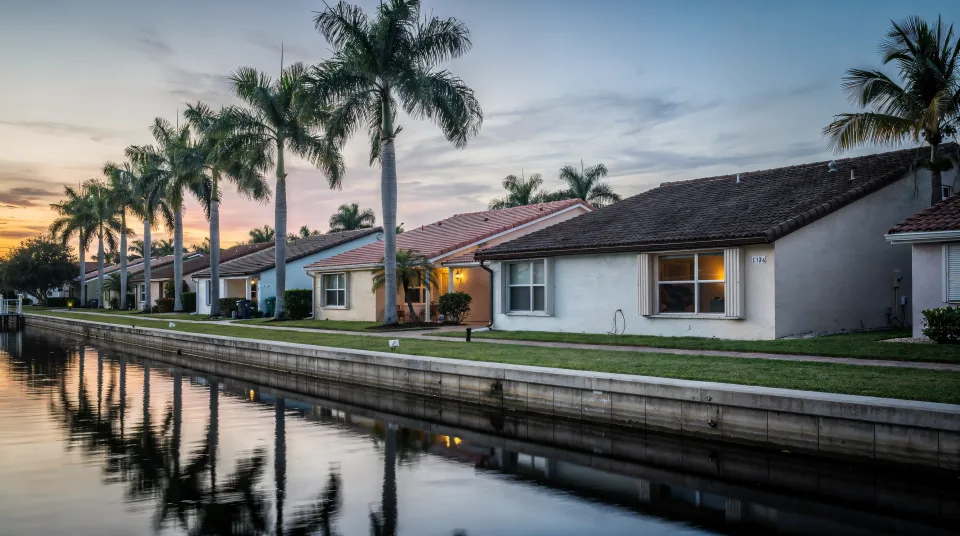Quiet canal-front street in Broward County at dusk, single-story 1970s concrete-block homes with barrel-tile roofs, a low seawall and a row of royal palms reflected in the water
