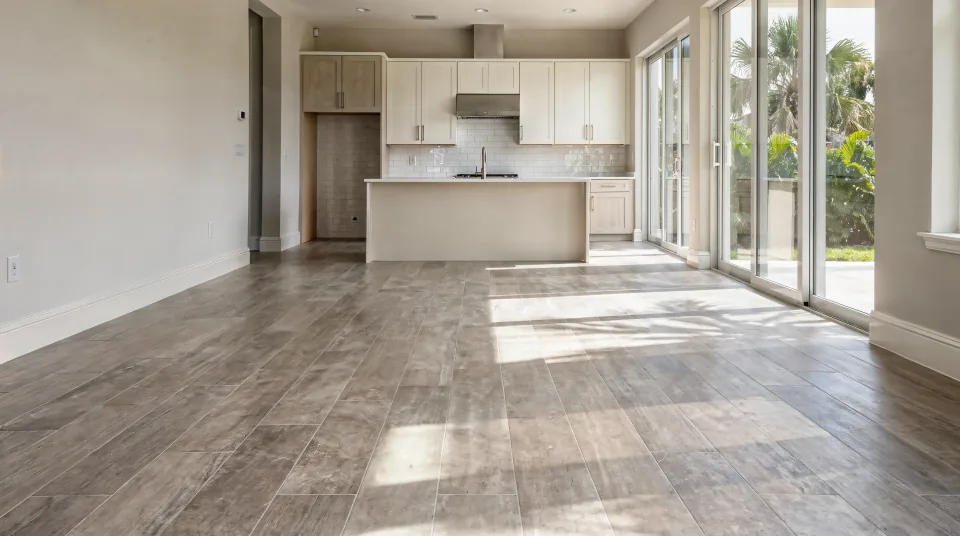 Open living room with large-format greige porcelain plank flooring running toward a kitchen with a white tiled backsplash, afternoon light from sliding glass doors