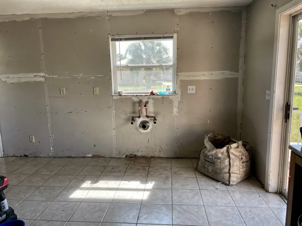 Kitchen with the old cabinets and counters removed, bare drywall and capped plumbing under the window