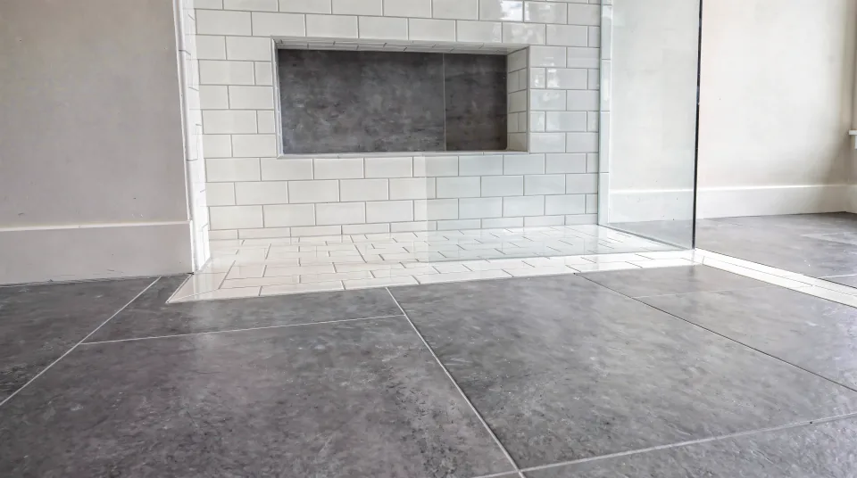 Finished bathroom floor in large-format grey porcelain with tight grout lines meeting a white subway-tile shower wall with a tiled niche