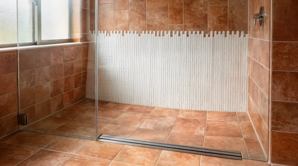 Walk-in shower in a one-story 1980s bathroom with terracotta-toned porcelain, a white picket-tile accent wall, a curbless entry with a linear drain and clear midday light