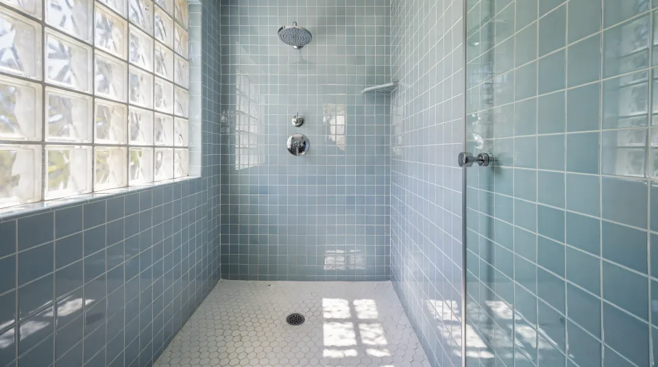 Walk-in shower in a 1950s bungalow bathroom with pale blue 4-inch square tile, a white hex floor, a chrome rain head and light through a glass-block window