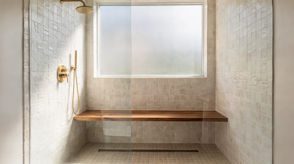 Walk-in shower built in a converted 60-inch alcove with warm white zellige-look tile, a teak bench, brushed brass fixtures and morning light through a frosted window