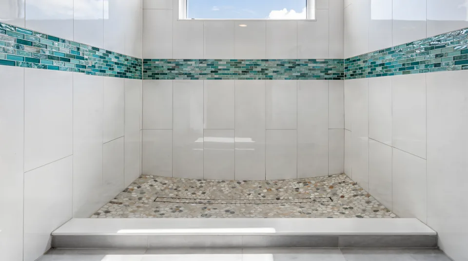Converted walk-in shower with an aqua glass mosaic accent strip, white large-format tile, a pebble floor, a low curb and bright coastal light from a high window