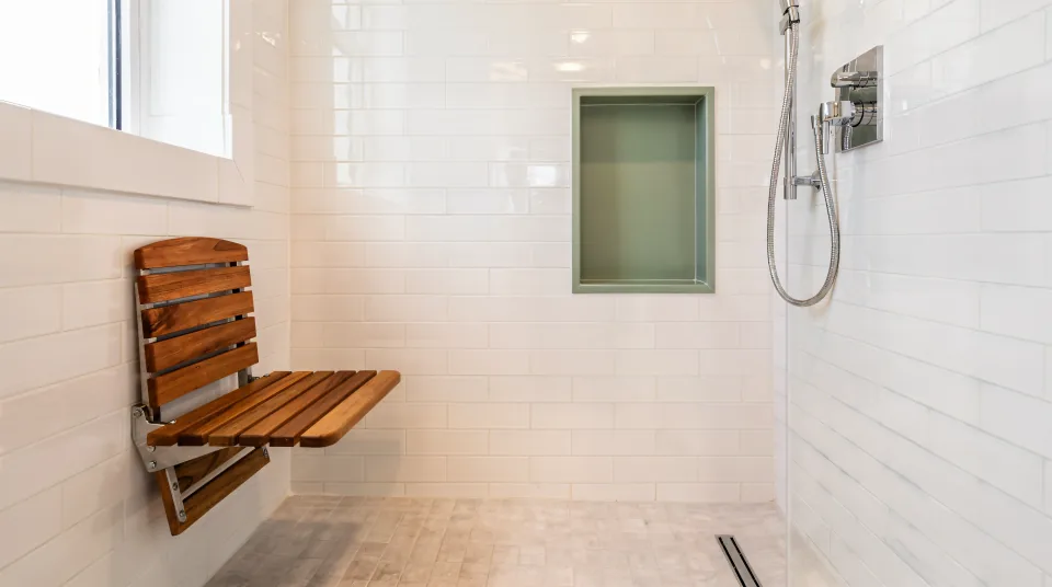 Compact condo walk-in shower in glossy white subway tile with a sage green tiled niche, a fold-down teak seat, a hand-held shower on a slide bar and bright afternoon light