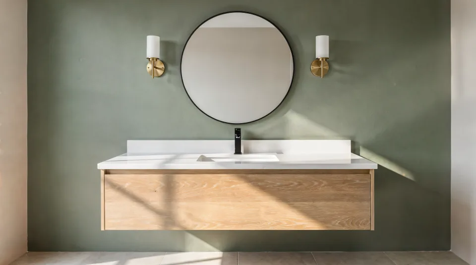 Floating white oak bathroom vanity with a quartz top, a matte black faucet, a round mirror and two brass sconces against a sage green wall