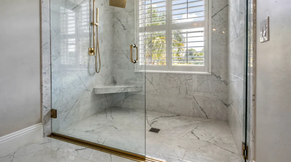 Finished curbless walk-in shower in a Boca Raton primary bath, honed white marble-look porcelain with grey veining, a wide brushed-brass linear drain at the threshold, a cantilevered bench, a hand shower on a slide bar and a frameless glass enclosure with a return panel, lit by a tall window with white plantation shutters