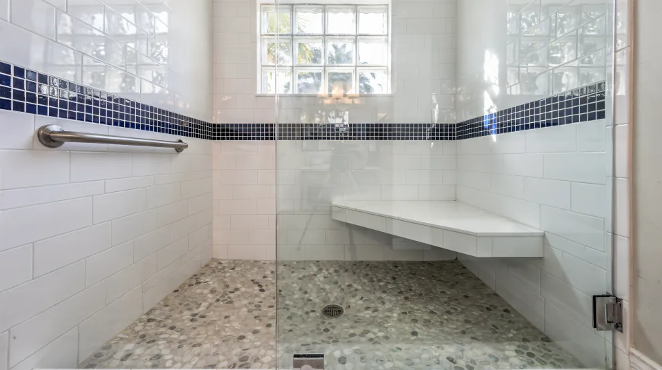 Finished curbless walk-in shower in a 1980s ranch bathroom, white 12-by-24 porcelain with a navy mosaic band at eye level, a pebble-look floor sloping to a center drain, a wall-mounted bench, a brushed nickel grab bar and a frameless glass panel lit by a glass-block window