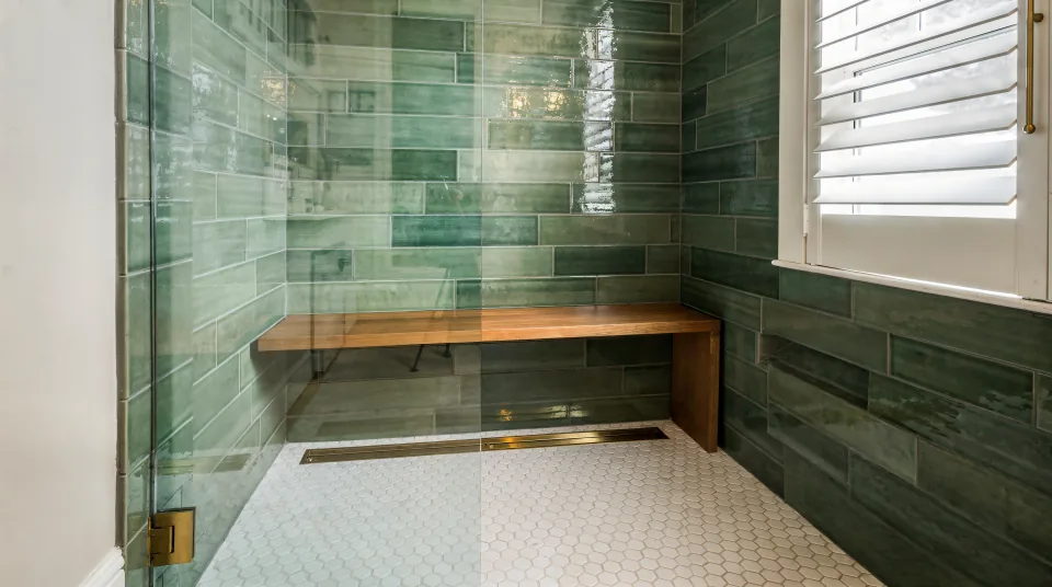 Finished curbless walk-in shower in a 1950s bungalow bathroom, green handmade-look wall tile, a white hexagon mosaic floor sloping to a brass linear drain, a teak bench and a frameless glass panel beside a louvered window