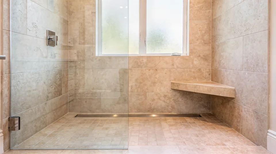 Finished curbless walk-in shower in warm sand-colored large-format porcelain with a long linear drain at the back wall, a tiled bench and a single frameless glass panel lit by a frosted window