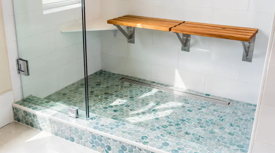 Finished low-threshold walk-in shower in a beach cottage bathroom, sea-glass blue penny-round mosaic on the shower floor rising to a shallow tiled curb, white large-format walls, a chrome linear drain, a fold-down teak seat and a clear glass panel in bright coastal daylight