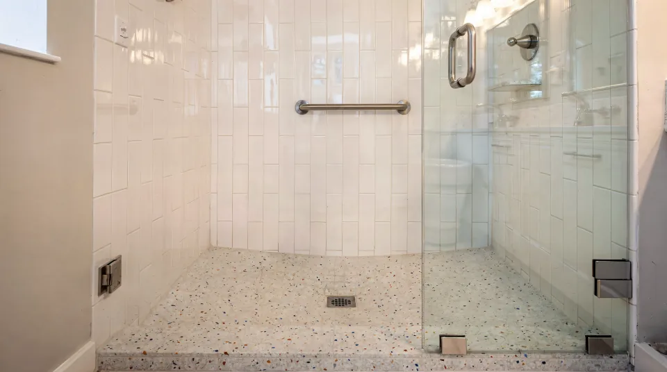 Finished compact walk-in shower in a condo bathroom, glossy white 3-by-12 tile laid vertically, a pale terrazzo-look floor sloping to a square center drain behind a low tiled curb, a brushed nickel grab bar and a clear fixed glass panel under warm vanity light