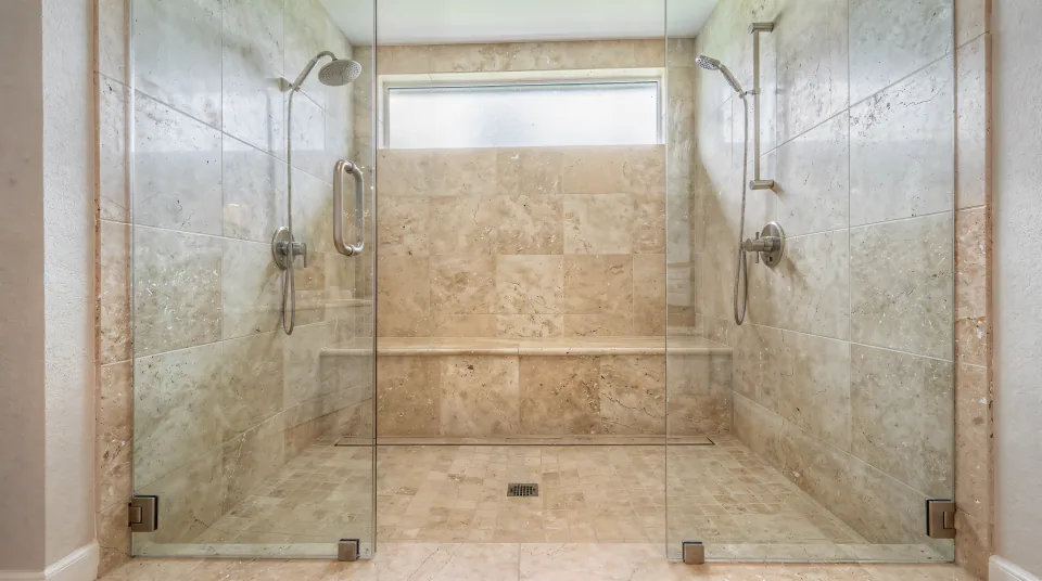 Finished zero-entry walk-in shower in a large primary bath, creamy travertine-look porcelain in 24-by-48 panels, a tile-top linear drain flush with the floor, a long built-in bench, a rain head and hand shower on a slide bar, and a floor-to-ceiling frameless glass wall with soft light from a frosted transom